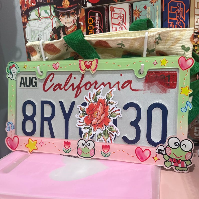 May include: A green and pink California license plate with the text "AUG California 8RY 30" and various decorative elements, including a flower, a music note, and a cartoon character.