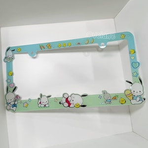 May include: A light blue license plate frame with cartoon characters and food illustrations. The frame has a green border with cartoon bunnies, chicks, and other graphics. The top of the frame has the text "@velaine".