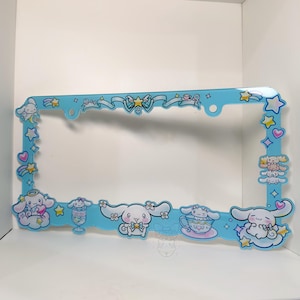 Puede incluir: Un marco de matr&iacute;cula azul claro con personajes de dibujos animados, estrellas, corazones y lazos. El marco tiene forma rectangular con esquinas redondeadas y est&aacute; decorado con varias figuras de dibujos animados.