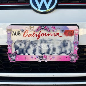 May include: A pink and purple license plate frame with cartoon characters and the word "California" in red. The frame has a decorative border with bows and hearts. The license plate is mounted on a white car.