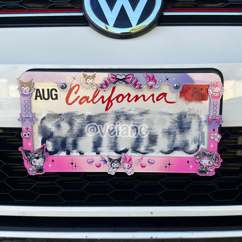 May include: A pink and purple license plate frame with cartoon characters and the word "California" in red. The frame has a decorative border with bows and hearts. The license plate is mounted on a white car.
