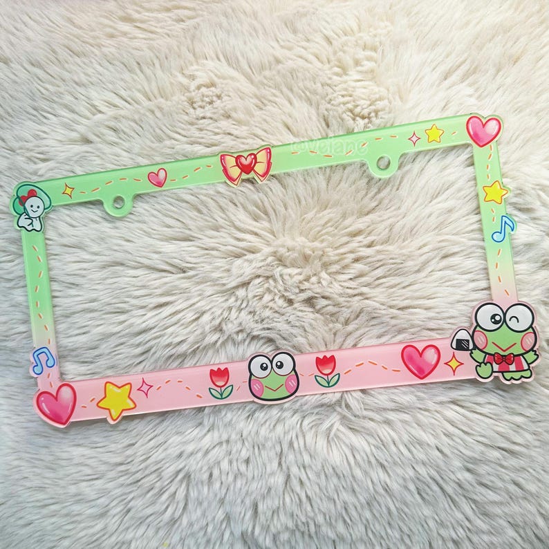 May include: A green and pink plastic license plate frame with a cartoon frog design. The frame has a variety of small cartoon details, including hearts, stars, musical notes, and flowers.