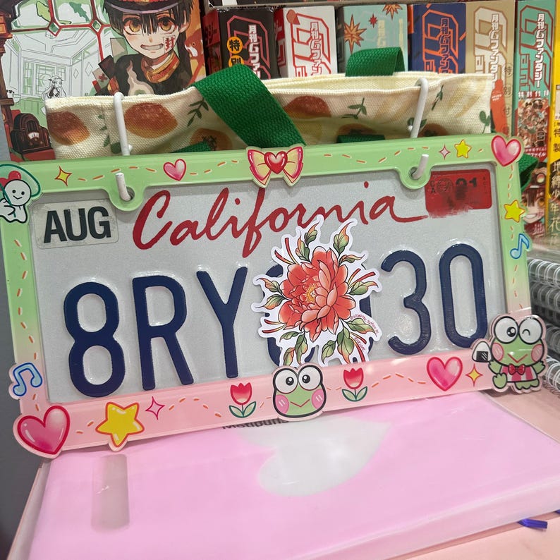 May include: A green and pink license plate frame with a California license plate that says "AUG 8RY 30". The frame is decorated with stickers of flowers, frogs, stars, and hearts.