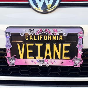 May include: A license plate with the word "VEIANE" in yellow letters. The license plate is framed with a pink and purple border, decorated with cartoon characters and bows. The word "CALIFORNIA" is printed at the top.