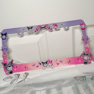 May include: A license plate frame with a gradient pink and purple design. The frame is decorated with cartoon characters, hearts, bows, and stars. The frame has a glossy finish and is designed for a car.
