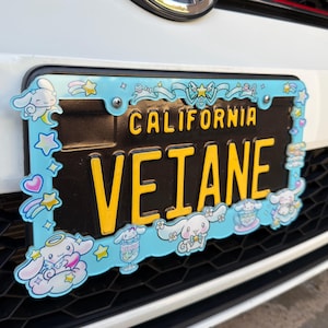 Puede incluir: Una placa de matr&iacute;cula negra de California con letras amarillas que dicen "VETANE". Est&aacute; enmarcada por un borde azul claro con personajes de dibujos animados, estrellas y corazones. La placa est&aacute; montada en un coche blanco.