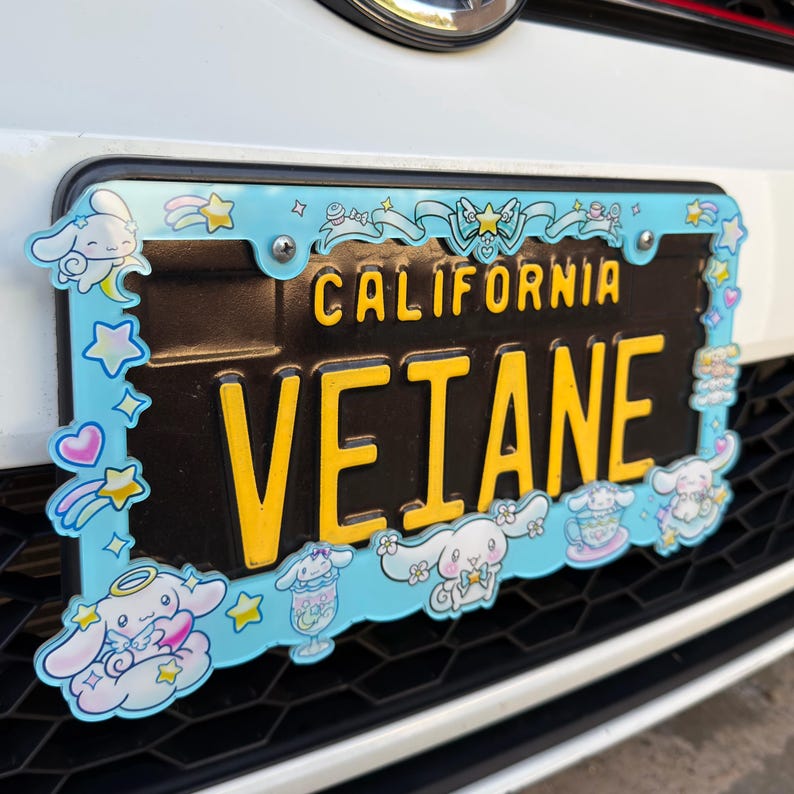 Puede incluir: Una placa de matr&iacute;cula negra de California con letras amarillas que dicen "VETANE". Est&aacute; enmarcada por un borde azul claro con personajes de dibujos animados, estrellas y corazones. La placa est&aacute; montada en un coche blanco.