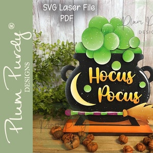 May include: A black cauldron with a green bubbling potion and a crescent moon design. The cauldron has the words "Hocus Pocus" written on it in gold letters. A wooden broom is attached to the side of the cauldron. The cauldron is sitting on a wooden base.