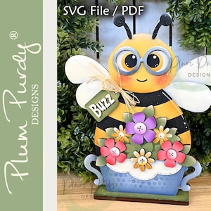 Può includere: Una stravagante decorazione in legno a tema ape. L'ape ha un corpo giallo con strisce nere, grandi occhi e indossa occhiali. Tiene un'etichetta con la scritta "Buzz". L'ape è decorata con fiori colorati e si trova in una fioriera blu. Il design è etichettato "SVG File / PDF".