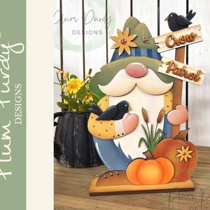 Könnte beinhalten: Eine Holzfigur eines Zwergs mit einem grünen Hut, einem gelben und braunen Hemd und einer blau-weiß karierten Weste. Der Zwerg hält einen schwarzen Vogel in seiner rechten Hand. Der Zwerg steht auf einem Holzsockel mit einem Schild, auf dem "Crow Patrol" steht. Vor dem Zwerg liegen zwei Kürbisse.