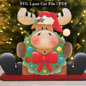 Puede incluir: Una decoración de madera cortada con láser con el texto SVG Laser Cut File / PDF. El alce lleva un gorro de Papá Noel rojo, sostiene una corona verde con adornos coloridos y un gran lazo rojo, y se asienta sobre una base roja con pies negros.