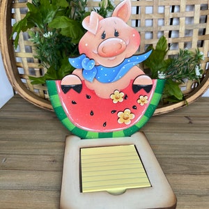 May include: A wooden pig figurine wearing a blue bandana sits in a red watermelon slice. The watermelon slice is green on the outside and has black seeds. The pig is holding the watermelon slice with its front legs. The figurine is attached to a wooden base with a yellow notepad.