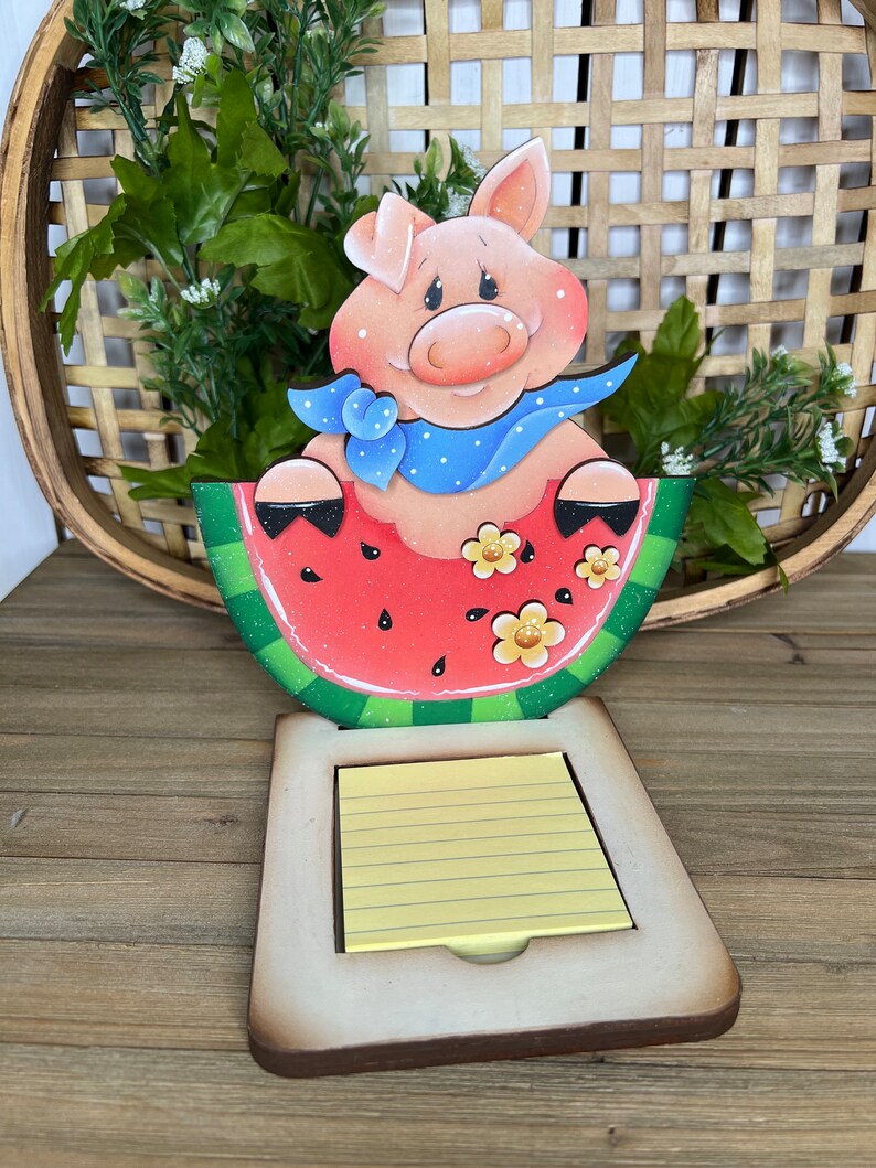 May include: A wooden pig figurine wearing a blue bandana sits in a red watermelon slice. The watermelon slice is green on the outside and has black seeds. The pig is holding the watermelon slice with its front legs. The figurine is attached to a wooden base with a yellow notepad.