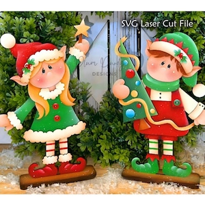 May include: Two flat wooden elf cutouts standing on rectangular bases. The left elf has blonde hair, a red hat, and a green dress. The right elf has brown hair, a green hat, and a red outfit holding a decorated tree. Text reads SVG Laser Cut File.