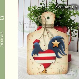 以下が含まれることがあります： 赤、白、青の雄鶏のデザインが施された木製のカッティングボード。雄鶏の尾羽には金色の星が飾られています。ボードはベージュの背景に白い星で飾られています。右下に「Plum Purdy Designs」の文字が見えます。