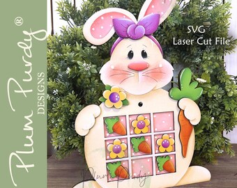 May include: Wooden Easter bunny decoration with a pink bow, polka-dotted ears, and a carrot. The bunny holds a carrot and has a grid of squares with carrots and flowers. The text "SVG Laser Cut File" is visible.