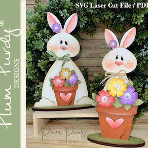Op de afbeelding: Twee houten paashaasdecoraties. De konijnen zijn wit met roze oren en wangen. Elk konijn houdt een bloempot met kleurrijke bloemen vast. De bloempotten hebben een hartvormige uitsparing. De tekst "SVG Laser Cut File / PDF" staat bovenaan.