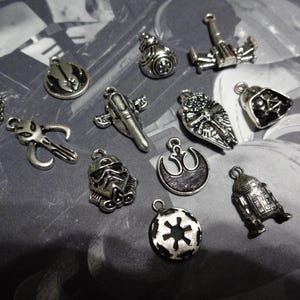 May include: A collection of silver-toned charms featuring iconic symbols and characters from a science fiction franchise. The charms include a helmet, a spaceship, a droid, and other recognizable shapes, all with a metallic finish.