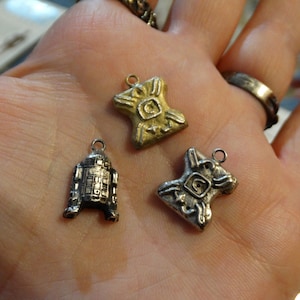 May include: Three silver and gold charms, one shaped like a robot, and two shaped like a stylized human figure. The charms are on a hand.