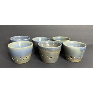 May include: A set of five ceramic bowls with a blue and tan glazed finish. Each bowl features a series of small, circular perforations arranged near the base, creating a distinct pattern around the lower exterior of the vessels.