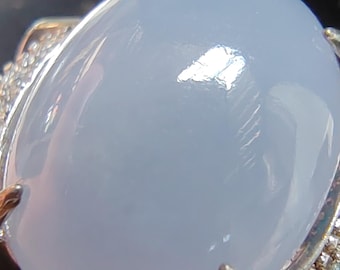 May include: A close-up of a silver ring featuring a large, oval-shaped, pale blue gemstone. The gemstone has a smooth, polished surface. The ring's band is adorned with small, clear gemstones.