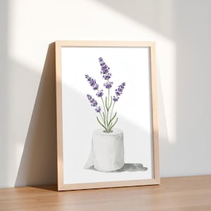 May include: A framed watercolor print featuring a roll of white toilet paper with lavender sprigs. The lavender has purple flowers and green stems. The print is in a light wood frame and is displayed on a wooden surface.