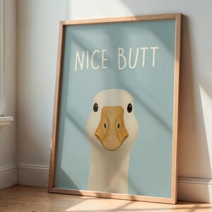 May include: A framed art print featuring a cartoon goose with the text "NICE BUTT" in white on a light blue background. The goose has a white head, a tan beak, and black eyes. The frame is made of light-colored wood.