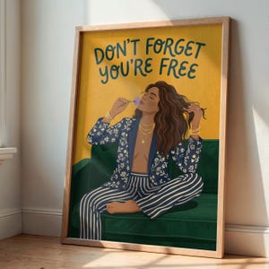 Könnte beinhalten: Gerahmter Kunstdruck mit dem Text "DON'T FORGET YOU'RE FREE". Das Kunstwerk zeigt eine Frau in einem blauen Blumenblazer und gestreiften Hosen, die auf einem grünen Sofa sitzt. Der Hintergrund ist gelb und der Rahmen hell.