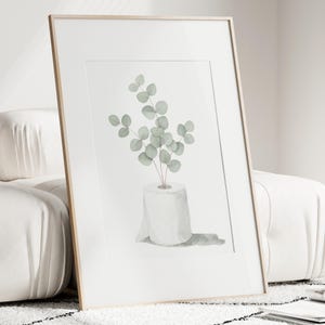 Op de afbeelding: Ingelijste kunstdruk. Waterverfillustratie van een rol toiletpapier met eucalyptusbladeren. Het kunstwerk heeft een minimalistische stijl en gebruikt zachte groene en grijze tinten tegen een witte achtergrond.