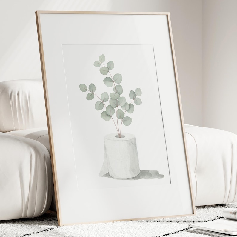 Op de afbeelding: Ingelijste kunstdruk. Waterverfillustratie van een rol toiletpapier met eucalyptusbladeren. Het kunstwerk heeft een minimalistische stijl en gebruikt zachte groene en grijze tinten tegen een witte achtergrond.