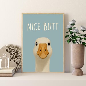 May include: A framed art print featuring a close-up of a white goose with a yellow beak and the text "NICE BUTT" on a light blue background. The artwork is displayed on a white shelf with decorative items and a vase with white flowers.