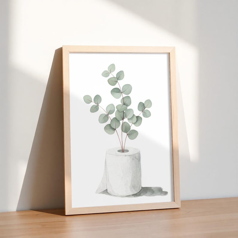 Op de afbeelding: Een ingelijste kunstdruk met een aquarel illustratie van eucalyptus takken in een toiletrol. Het kunstwerk heeft een licht houten frame en staat tegen een witte achtergrond. De eucalyptusbladeren zijn groen, het toiletpapier is wit.
