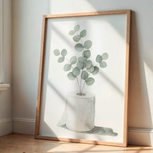 Op de afbeelding: Een ingelijste aquarelprint met een rol toiletpapier met eucalyptus takken. Het kunstwerk heeft een minimalistische stijl met zachte groene en witte tinten. De lijst is gemaakt van lichtgekleurd hout en de print leunt tegen een witte muur.