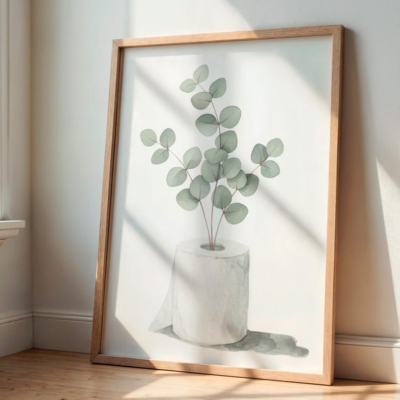 Op de afbeelding: Een ingelijste aquarelprint met een rol toiletpapier met eucalyptus takken. Het kunstwerk heeft een minimalistische stijl met zachte groene en witte tinten. De lijst is gemaakt van lichtgekleurd hout en de print leunt tegen een witte muur.