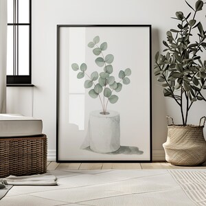 Op de afbeelding: Een ingelijste kunstdruk met een aquarel illustratie van eucalyptus takken die uit een rol toiletpapier komen. Het kunstwerk heeft een witte achtergrond en een zwarte lijst. De print wordt getoond in een modern, minimalistisch interieur.