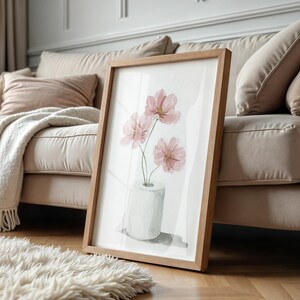 Puede incluir: Una impresi&oacute;n art&iacute;stica enmarcada con una ilustraci&oacute;n en acuarela de flores de cosmos rosas en un jarr&oacute;n blanco. La obra de arte est&aacute; sobre un fondo blanco y enmarcada en madera marr&oacute;n claro. La impresi&oacute;n est&aacute; apoyada contra un sof&aacute; beige y una alfombra blanca y esponjosa.