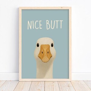 May include: Framed art print featuring a close-up illustration of a white goose with a yellow beak and black eyes. The text "NICE BUTT" is written in white above the goose on a light blue background. The frame is made of light wood.