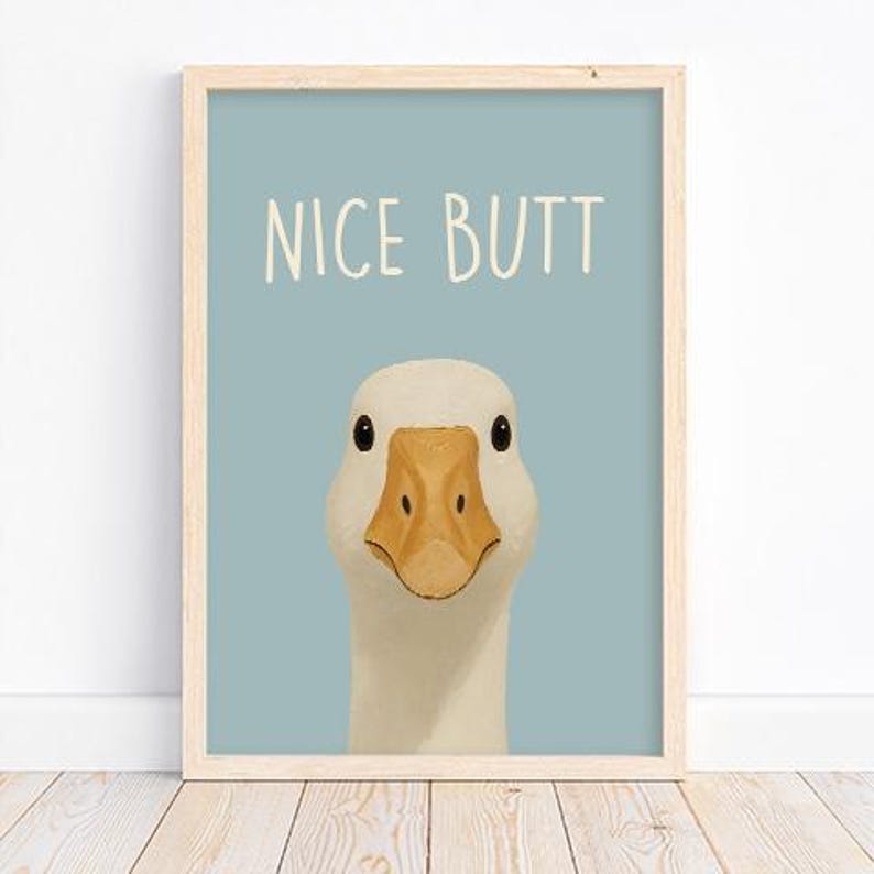 May include: Framed art print featuring a close-up illustration of a white goose with a yellow beak and black eyes. The text "NICE BUTT" is written in white above the goose on a light blue background. The frame is made of light wood.