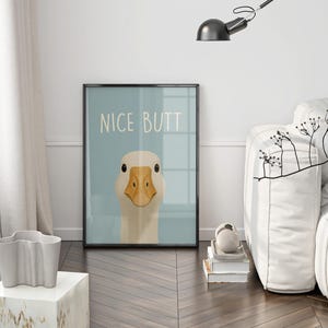 May include: Framed art print featuring a cartoon goose with a light blue background. The print has the words "NICE BUTT" in white. The goose has a beige head and a yellow beak. The print is in a black frame.