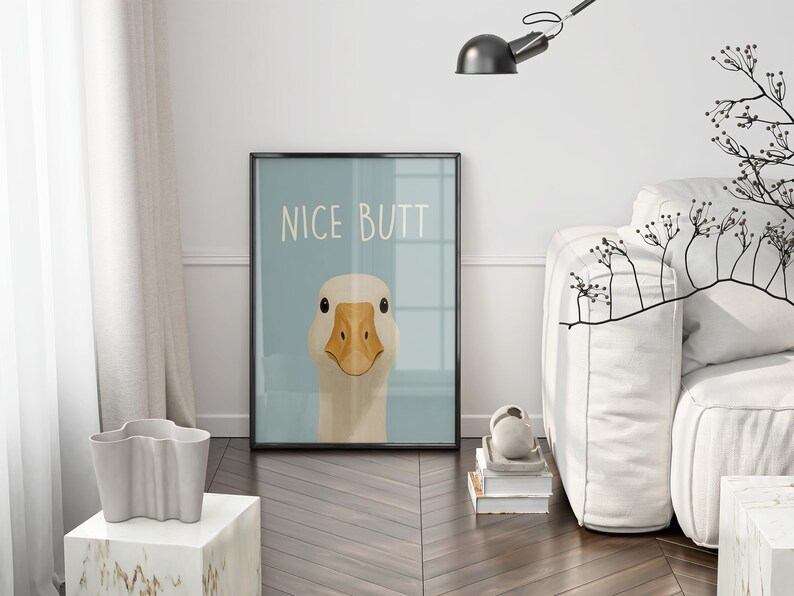 May include: Framed art print featuring a cartoon goose with a light blue background. The print has the words "NICE BUTT" in white. The goose has a beige head and a yellow beak. The print is in a black frame.