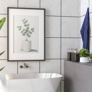 Op de afbeelding: Een ingelijste aquarelprint van eucalyptus takjes in een rol toiletpapier. Het kunstwerk staat in een zwarte lijst en hangt aan een wit betegelde muur. Een witte badkuip en badkameraccessoires staan op de voorgrond.