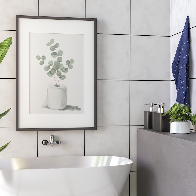 Op de afbeelding: Een ingelijste aquarelprint van eucalyptus takjes in een rol toiletpapier. Het kunstwerk staat in een zwarte lijst en hangt aan een wit betegelde muur. Een witte badkuip en badkameraccessoires staan op de voorgrond.