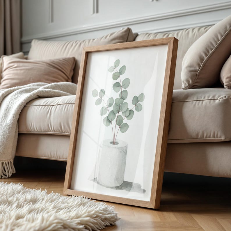 Op de afbeelding: Een ingelijste botanische print met een aquarel illustratie van eucalyptusbladeren in een witte vaas. Het kunstwerk staat in een lichtbruine houten lijst en leunt tegen een beige bank. De afbeelding staat op een houten vloer met een wit, pluizig tapijt.