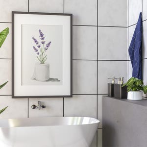 May include: Framed watercolor print featuring lavender sprigs in a toilet paper roll. The artwork has a white background and is set in a dark gray frame. The image is displayed in a bathroom setting with a white tub and gray tile.