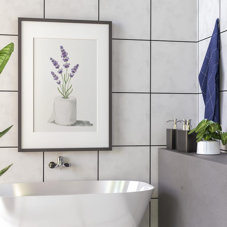 May include: Framed watercolor print featuring lavender sprigs in a toilet paper roll. The artwork has a white background and is set in a dark gray frame. The image is displayed in a bathroom setting with a white tub and gray tile.