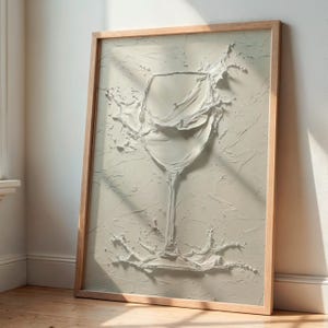 Könnte beinhalten: Ein gerahmtes Kunstwerk mit einer strukturierten, monochromen Darstellung eines Weinglases mit herausspritzender Flüssigkeit. Das Kunstwerk ist in einer hellen, neutralen Farbe gehalten und befindet sich in einem Naturholzrahmen, der an einer weißen Wand lehnt.