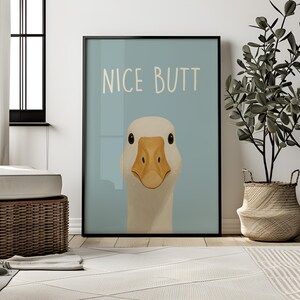 May include: Framed art print featuring a close-up illustration of a white goose with a yellow beak and the text "NICE BUTT" on a light blue background. The artwork is displayed in a black frame.
