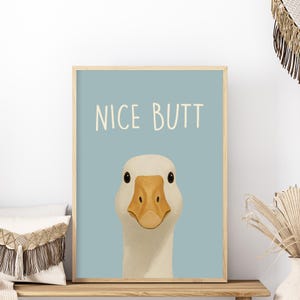 May include: A framed print featuring a cartoon goose with the text "NICE BUTT" in a playful font. The goose has white feathers, a yellow beak, and black eyes, set against a light blue background. The frame is a light wood color.