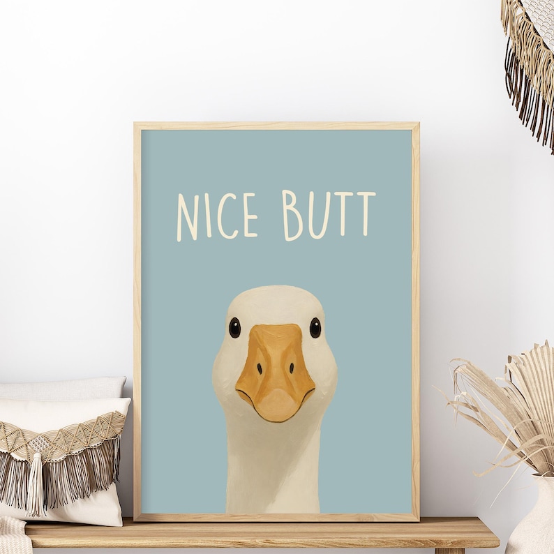 May include: A framed print featuring a cartoon goose with the text "NICE BUTT" in a playful font. The goose has white feathers, a yellow beak, and black eyes, set against a light blue background. The frame is a light wood color.