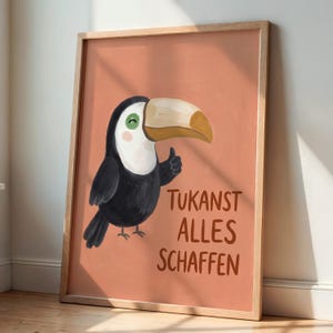 Könnte beinhalten: Ein gerahmter Kunstdruck mit einem Cartoon-Tukan mit großem beigen Schnabel, schwarzem Körper und weißer Brust auf terrakottafarbenem Hintergrund. Der Tukan zeigt einen Daumen hoch. Der Text TUKANST ALLES SCHAFFEN steht braun unter dem Vogel.